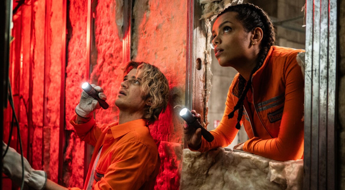 Auf dem Bild zur Cold Storage Filmkritik sind Naomi Williams, gespielt von Georgina Campbell, und Travis „Teacake“ Meacham, dargestellt von Joe Keery, in einer spannungsgeladenen Filmszene zu sehen. Beide tragen orange Arbeitskleidung und leuchten mit Taschenlampen in einen dunklen, industriellen Raum. Die rote Beleuchtung und ihre konzentrierten Blicke erzeugen eine bedrohliche, nervöse Atmosphäre.