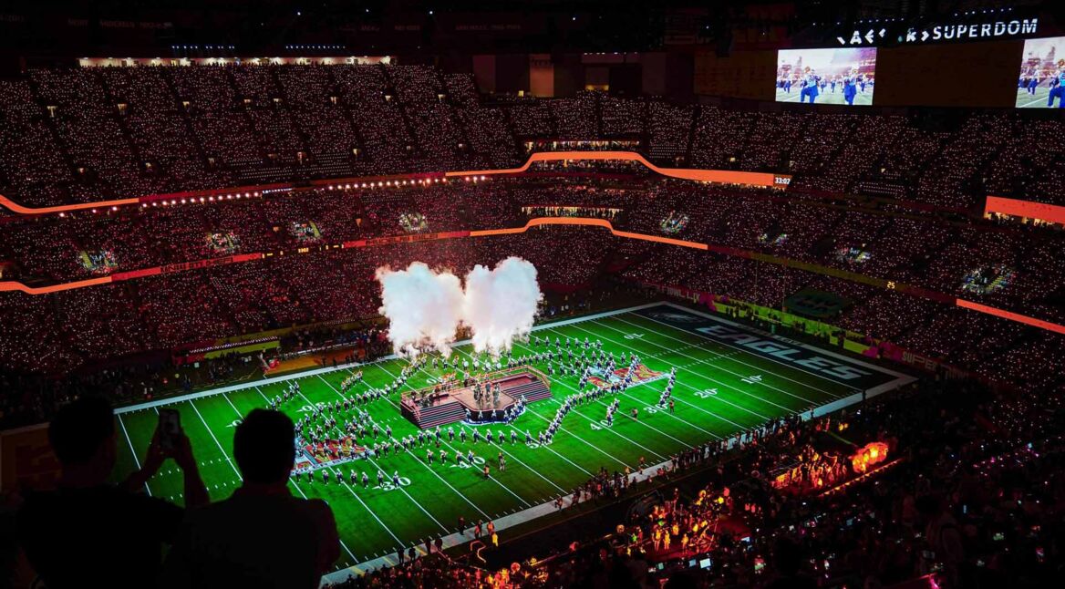 Auf dem Bild in den GigaTV-Highlights im Februar 2026 siehst Du eine Szene vom Super Bowl. In einem riesigen Stadion sind tausende Zuschauer zu sehen, während auf dem Spielfeld eine spektakuläre Halbzeitshow mit Rauch, Licht und Choreografien stattfindet. Die Stimmung wirkt energiegeladen, monumental und voller sportlicher und musikalischer Highlights.