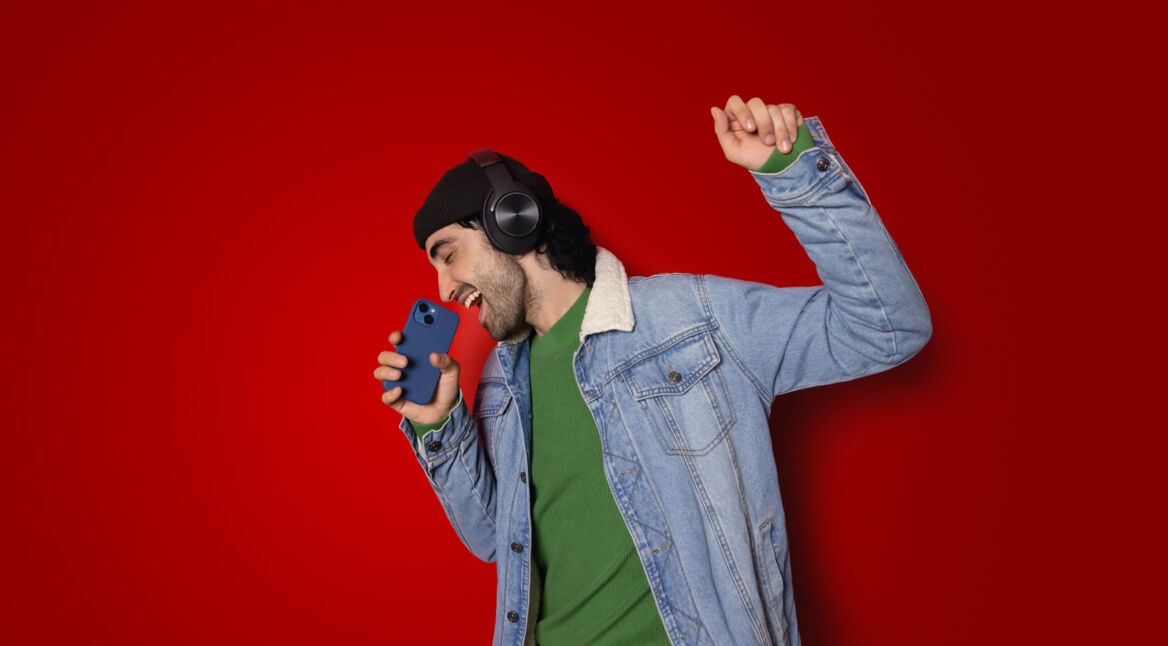 Auf dem Bild in der Musik-Streamingdienste-Übersicht ist ein junger Mann vor rotem Hintergrund zu sehen, der mit geschlossenen Augen begeistert in ein Smartphone singt. Er trägt schwarze Over-Ear-Kopfhörer, eine schwarze Mütze, ein grünes Shirt und eine Jeansjacke. Seine erhobene Faust unterstreicht die ausgelassene, energiegeladene Stimmung beim Musikhören.