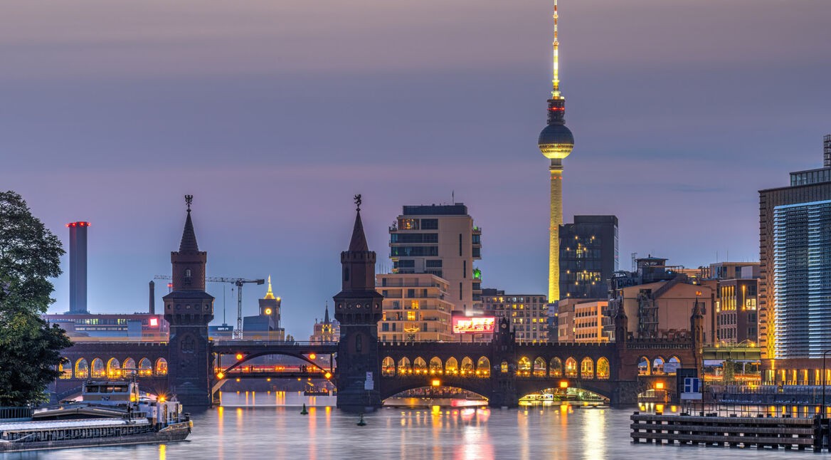 Blick auf die Berliner Skyline bei Dämmerung mit beleuchteter Oberbaumbrücke im Vordergrund und dem Fernsehturm im Hintergrund.