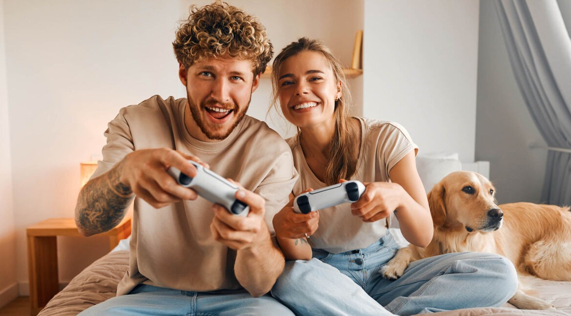 Ein junges Paar sitzt lachend auf einem Bett und spielt mit zwei Gamecontrollern ein Videospiel, während ein Golden Retriever entspannt neben ihnen liegt.