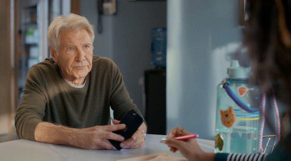 Harrison Ford sitzt an einem Tisch. Er ist frontal zu sehen und hält ein Smartphone in der Hand. Er spricht mit einer nicht erkennbaren Person, deren Hand einen roten Stift hält.