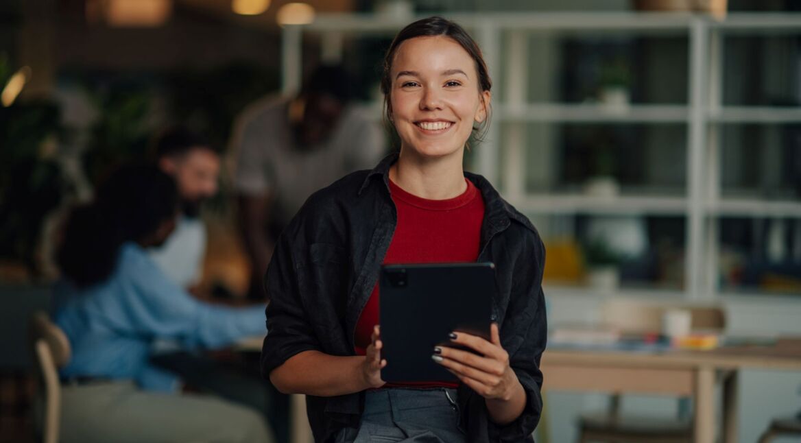 Young businesswoman holding tablet and smiling in modern office with colleagues working in background