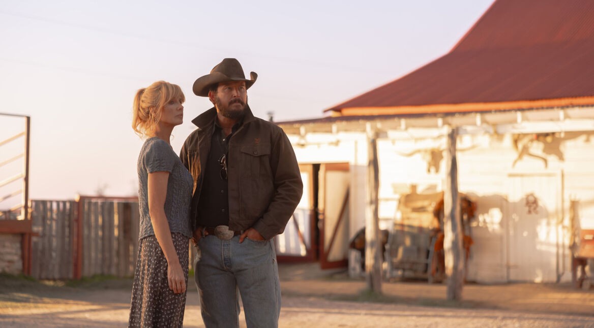 Auf dem Bild zur Dutton Ranch stehen Kelly Reilly als Beth Dutton und Cole Hauser als Rip Wheeler nebeneinander vor einer Ranch im warmen Abendlicht. Beth trägt ein schlichtes Kleid und wirkt nachdenklich, während Rip mit Cowboyhut und ernster Miene in die Ferne blickt. Die Szene vermittelt eine ruhige, zugleich spannungsgeladene Western-Atmosphäre.