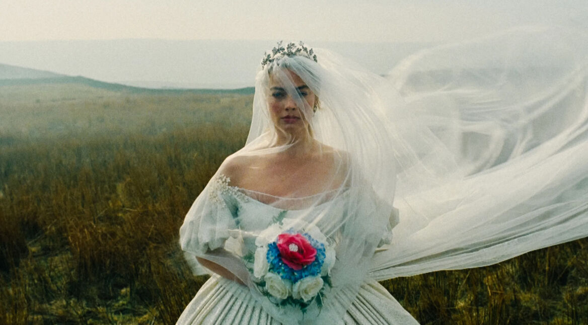 Auf dem Bild in den GigaTV-Highlights im April 2026 siehst Du eine Szene aus „Wuthering Heights – Sturmhöhe“ mit Catherine, gespielt von Margot Robbie. Sie steht in einer weiten, windigen Landschaft in einem weißen Kleid mit Schleier. Ihr ernster Blick und die düstere Naturkulisse erzeugen eine melancholische, intensive und romantisch-dramatische Stimmung.