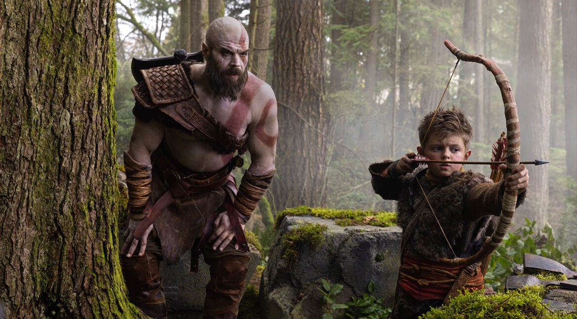 Auf dem Bild aus God of War sind Kratos und Atreus in einem dichten Wald zu sehen. Links steht Kratos, ein großer muskulöser Krieger mit Glatze, roter Kriegsbemalung und Lederrüstung, der aufmerksam beobachtet. Rechts spannt Atreus konzentriert seinen Bogen mit eingelegtem Pfeil. Die Szene wirkt angespannt und lehrreich, während Kratos den jungen Jäger im Wald trainiert.