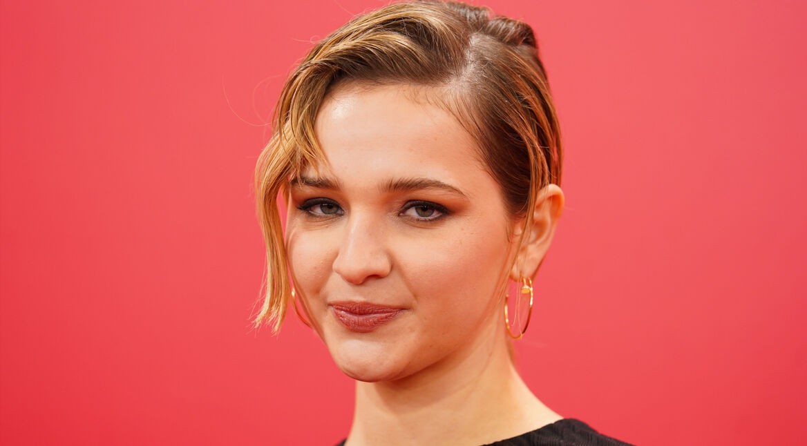 Auf diesem Porträt zur möglichen Struwwelpeter-Serie ist die Schauspielerin Lisa Vicari vor einem kräftig roten Hintergrund zu sehen. Sie trägt ein schwarzes Oberteil und goldene Creolen. Ihr kurzes blondes Haar ist seitlich gestylt, eine Strähne fällt locker ins Gesicht. Ihr Blick wirkt ruhig und selbstbewusst, wodurch die Nahaufnahme eine konzentrierte und leicht nachdenkliche Stimmung erzeugt.