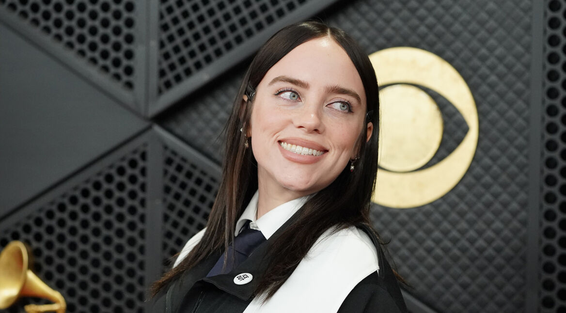 Auf diesem Bild ist Billie Eilish bei einem Event zu sehen. Sie trägt ein schwarz-weißes Outfit mit Hemd, Krawatte und Jacke und blickt mit einem freundlichen Lächeln leicht nach links. Hinter ihr befindet sich ein großes goldenes Grammophon-Symbol vor einer dunklen, strukturierten Wand. Das Bild wird symbolisch für den The Bell Jar-Film verwendet.