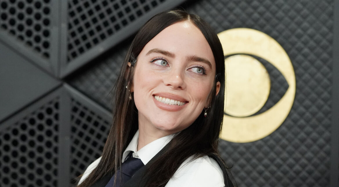 Auf diesem Bild ist Billie Eilish bei einem Event zu sehen. Sie trägt ein schwarz-weißes Outfit mit Hemd, Krawatte und Jacke und blickt mit einem freundlichen Lächeln leicht nach links. Hinter ihr befindet sich ein großes goldenes Grammophon-Symbol vor einer dunklen, strukturierten Wand. Das Bild wird symbolisch für den The Bell Jar-Film verwendet.