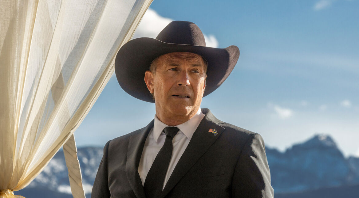 Auf dem Bild zur Yellowstone Serien-Reihenfolge ist John Dutton zu sehen, gespielt von Kevin Costner, der in einem schwarzen Anzug mit Krawatte und Cowboyhut vor einer beeindruckenden Berglandschaft steht. Sein Blick wirkt ernst und nachdenklich, während ein heller Vorhang neben ihm weht. Die Szene vermittelt eine ruhige, aber zugleich würdevolle und leicht angespannte Stimmung.