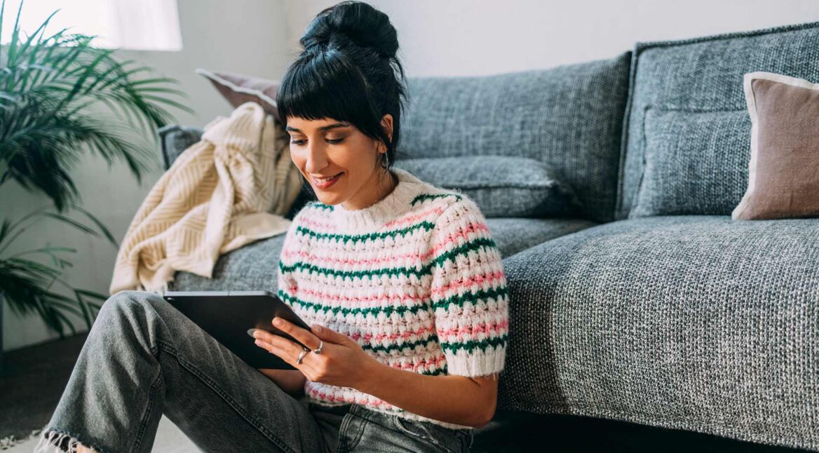 Auf diesem Bild zum Thema Tablet ohne SIM-Karte nutzen sitzt eine junge Frau entspannt auf dem Boden vor einem grauen Sofa in einem gemütlichen Wohnzimmer. Sie hält ein Tablet in den Händen und lächelt beim Lesen.