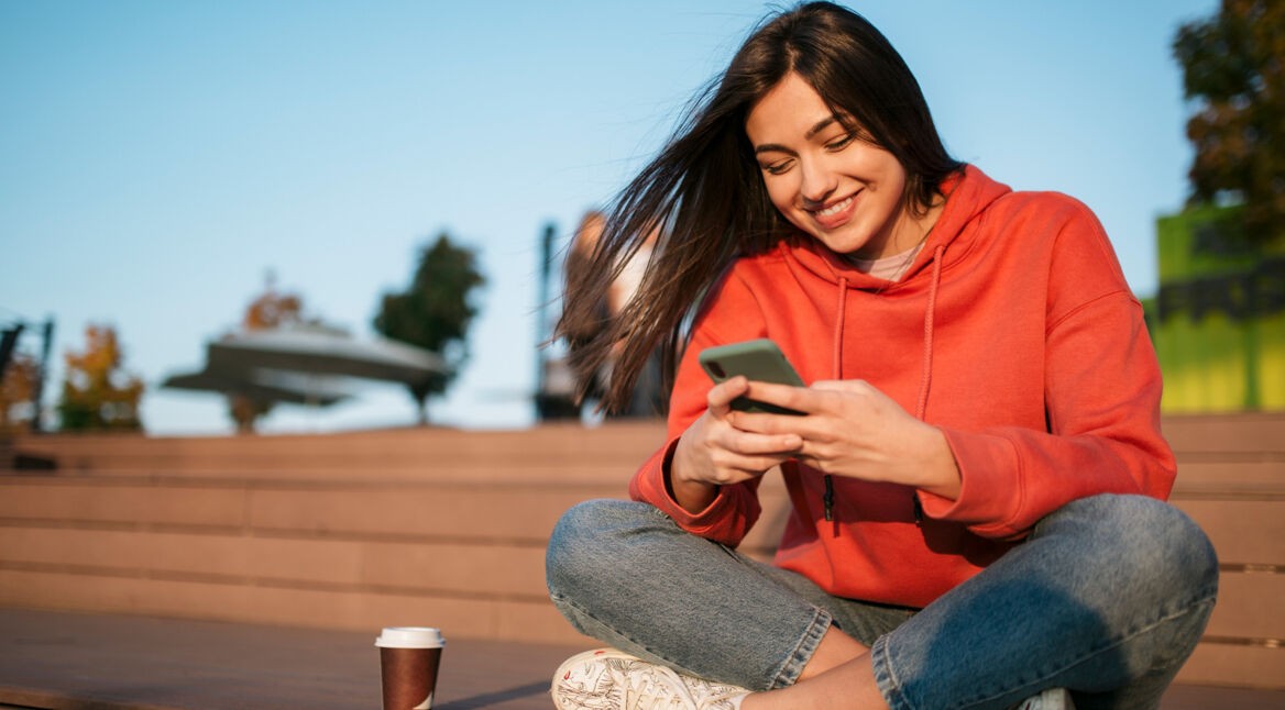 Auf dem Bild zum Thema Datenvolumen sparen sitzt eine junge Frau im roten Hoodie entspannt auf einer Holzbank im Freien. Sie lächelt zufrieden, während sie auf ihr Smartphone schaut. Neben ihr steht ein Coffee-to-go-Becher. Die warme Sonne, der blaue Himmel und die lockere Haltung erzeugen eine freundliche, unbeschwerte Stimmung.