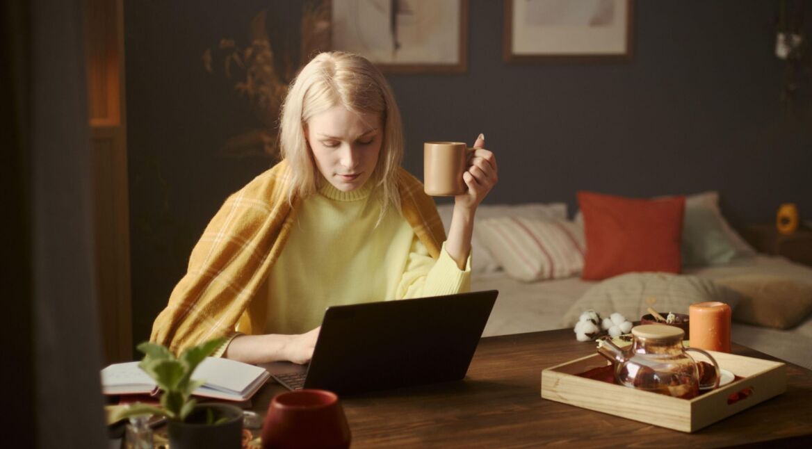 Junge Frau sitzt im Wohnzimmer an einem Tisch und arbeitet an ihrem Laptop.