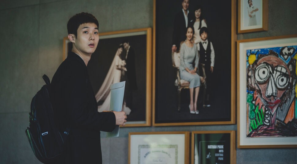 Ki-woo steht in Parasite vor einer Wand mit Familienfotos im Haus der Familie Park. Foto: Koch Films / Capelight Pictures