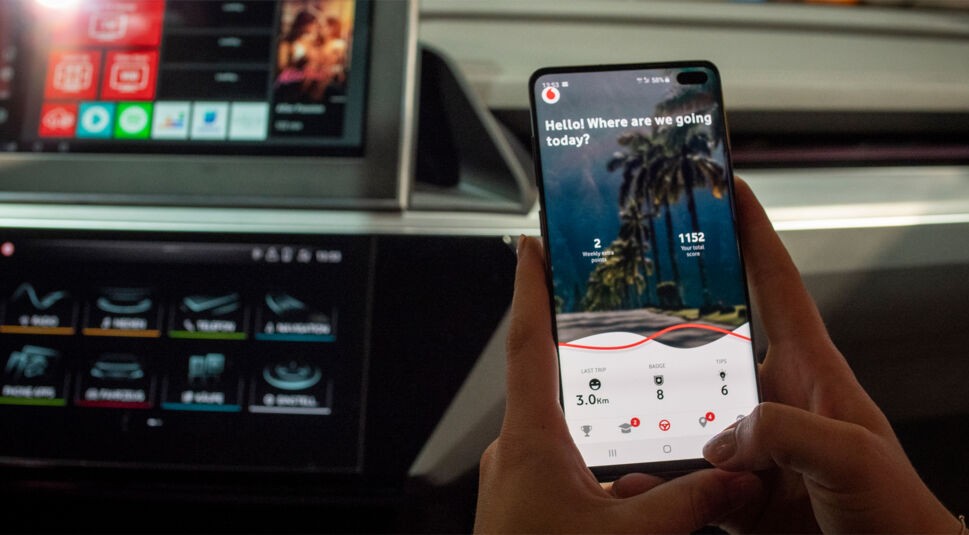 Hand hält Handy mit geöffneter App von Vodafone und EMIL in Fahrzeug-Cockpit.