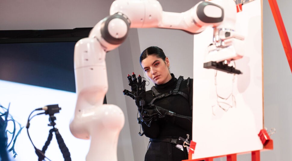 Die Künstlerin Shev Lunatic zieht einen Motion-Capturing-Anzug an, im Vordergrund ein Roboter, der ein Portrait zeichnet.