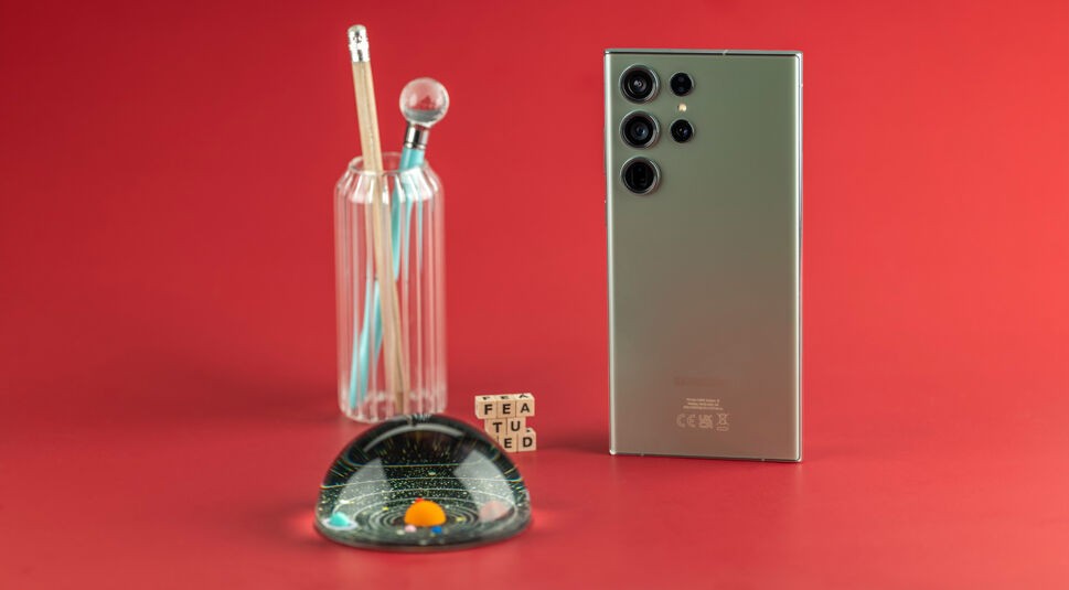 Smartphone von hinten stehend auf rotem Untergrund neben bunter Glaskugel, Stiftehalter und Buchstabenwürfeln