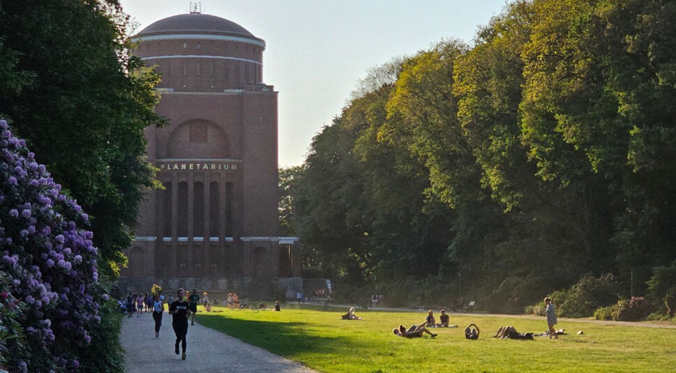 Ein mit Büschen und Bäumen gesäumter Weg mit einzelnen Personen, der auf einen roten Backsteinturm zuläuft, das Hamburger Planetarium