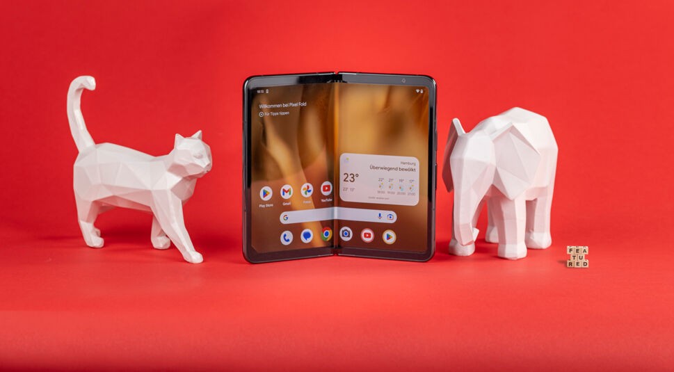 Falt-Smartphone Pixel Fold aufgeklappt vor rotem Hintergrund, eingerahmt von zwei weißen Tierfiguren