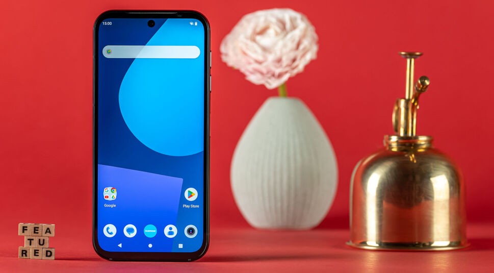 Das Fairphone 5 von vorne neben Buchstabenwürfeln, Blumenvase und einer Lötlampe vor rotem Hintergrund