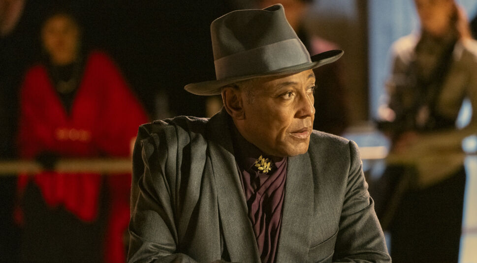 Auf dem Bild aus dem Film "Megalopolis" ist Giancarlo Esposito zu sehen. Er trägt einen eleganten grauen Anzug und einen passenden Fedora-Hut, während er zur Seite blickt. Sein Gesichtsausdruck ist ernst und nachdenklich, was auf eine bedeutungsvolle Szene hindeutet. Im Hintergrund sind unscharfe Gestalten zu erkennen, die eine geheimnisvolle und spannende Atmosphäre erzeugen.