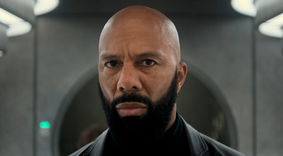 Auf diesem Bild aus Silo Staffel 2 ist der Schauspieler Common zu sehen. Er steht vor einem Hintergrund, der metallisch und industriell wirkt, passend zur dystopischen Umgebung der Serie. Common trägt einen schwarzen Mantel, der die ernste und dramatische Atmosphäre unterstreicht. Sein Blick ist intensiv und direkt in die Kamera gerichtet, was ein Gefühl von Entschlossenheit und Stärke vermittelt. Seine Gesichtszüge sind markant, mit einer Glatze und einem gepflegten Vollbart, die seinen charismatischen Ausdruck betonen. Der fokussierte Ausdruck in seinen Augen könnte auf eine kritische oder spannungsgeladene Szene hindeuten.