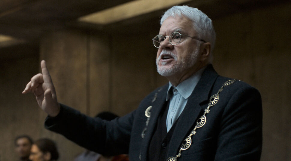 Auf dem Bild aus Silo Staffel 2 ist Tim Robbins zu sehen. Er trägt eine dunkle Jacke, die mit dekorativen goldfarbenen Knöpfen verziert ist, und ein graues Hemd. Robbins, mit seiner markanten weißen Haarpracht und Brille, hebt seine Hand mit ausgestrecktem Zeigefinger, was darauf hindeutet, dass er gerade eine bedeutsame oder argumentative Aussage macht. Der Hintergrund ist schlicht und wirkt wie der Innenraum einer rustikalen, holzverkleideten Struktur, was zu der dystopischen Atmosphäre der Serie passt.