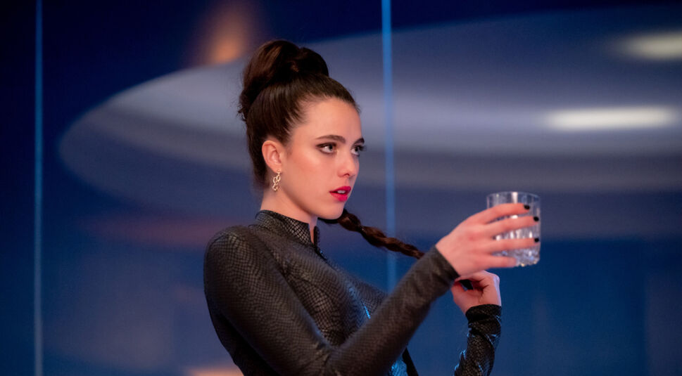 Im Bild aus dem Film The Substance ist Schauspielerin Margaret Qualley als Sue zu sehen. Eine junge, dunkelhaarige Frau. Sie schaut mit offenem Mund nach rechts und hält mit der rechten Hand ein Wasserglas vor sich. Mit der linken Hand spielt sie an ihren Haaren herum, die streng hochgebunden und in einem langen Zopf geflochen sind. Sie trägt einen schwarzen Gymnastikanzug und schwarzen Nagellack. Ihre Lippen sind in einem hellen Rot geschminkt und ihre Augen durch schwarzen Kayal betont.