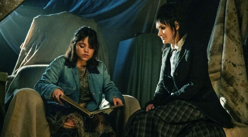 Auf dem Bild zu "Beetlejuice Beetlejuice Ende erklärt" sitzen Jenna Ortega als Astrid und Winona Ryder als Lydia nebeneinander. Astrid, in einer blauen Jacke, schaut konzentriert auf ein aufgeschlagenes Buch in ihren Händen, während Lydia in einem dunklen, karierten Outfit lächelnd neben ihr sitzt. Die Szene spielt sich in einem schwach beleuchteten Raum ab, umgeben von verhüllten Möbeln, was eine geheimnisvolle und leicht melancholische Atmosphäre erzeugt.