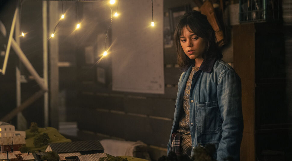 Auf dem Bild zu "Beetlejuice Beetlejuice Ende erklärt" ist Jenna Ortega als Astrid zu sehen, wie sie nachdenklich vor einem detailreichen Miniaturmodell einer Stadt steht. Sie trägt eine blaue Jacke und blickt ernst auf das Modell, während im Hintergrund warme Lichter in der dunklen, leicht chaotischen Umgebung hängen und für eine stimmungsvolle Beleuchtung sorgen. Die Szene vermittelt eine ruhige, nachdenkliche Atmosphäre und deutet auf einen wichtigen Moment im Film hin.