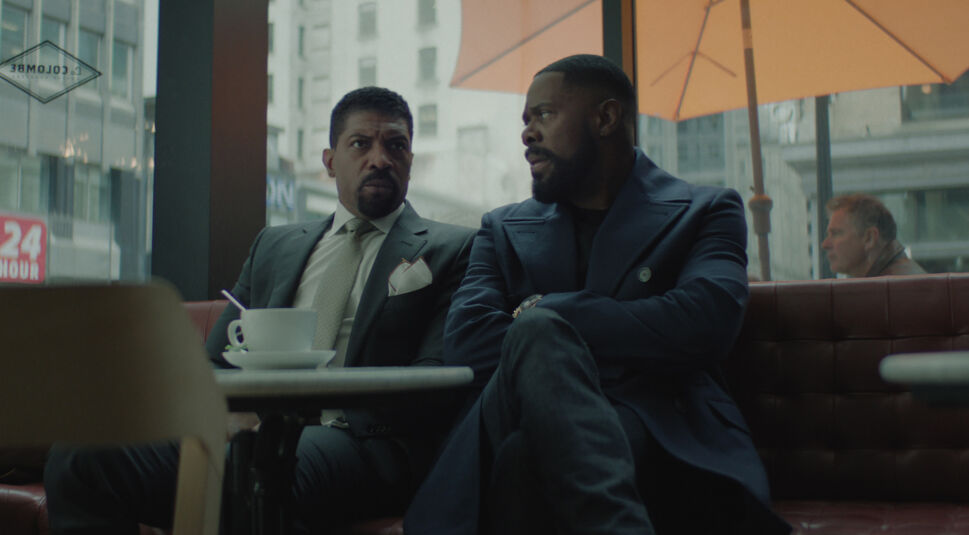 Auf dem Bild zur Serie The Madness sind Deon Cole als Kwesi Dupree und Colman Domingo als Muncie Daniels zu sehen. Die beiden sitzen in einem modernen Café an einem Tisch, Kwesi trägt einen eleganten Anzug mit Krawatte, während Muncie in einem dunkelblauen Mantel lässig gekleidet ist. Beide wirken in ein intensives Gespräch vertieft, während im Hintergrund eine urbane Szenerie mit großen Fenstern und einem orangefarbenen Sonnenschirm sichtbar ist.