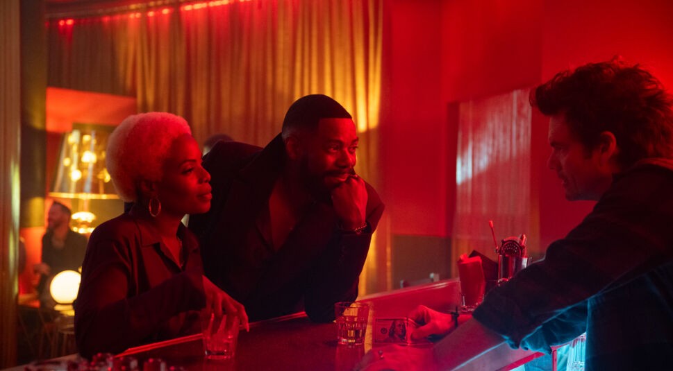 Auf dem Bild zur Serie The Madness sind Marsha Stephanie Blake als Elena und Colman Domingo als Muncie Daniels zu sehen. Beide stehen an einer Bar in einer stimmungsvoll rot beleuchteten Umgebung. Elena hält ein Glas in der Hand und blickt lächelnd auf den Barkeeper, während Muncie nachdenklich mit der Hand sein Kinn stützt. Der Barkeeper wirkt konzentriert, und die Szene vermittelt eine subtile Spannung und Vertrautheit.