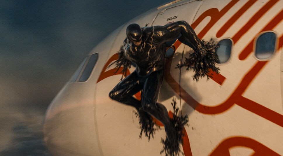 In dieser Szene aus Venom 3 sieht man Tom Hardy als Venom, wie er aus einem Flugzeug springt. Die schwarze, muskulöse Gestalt des Symbionten mit seinem markanten, bedrohlichen Grinsen haftet an der Außenwand des Flugzeugs, während er aus einer geöffneten Tür herausspringt. Die Szene wirkt actiongeladen und intensiv, mit einem düsteren Himmel im Hintergrund.