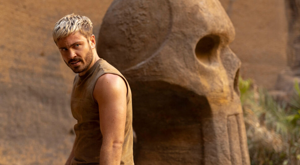 In „Die drei ??? – Toteninsel“ steht Olin, gespielt von Jannik Schümann, vor einer gigantischen Steinskulptur in Totenschädelform. Mit blondierten Haaren, ärmellosem Shirt und angespanntem Gesichtsausdruck wirkt er entschlossen und kampfbereit.