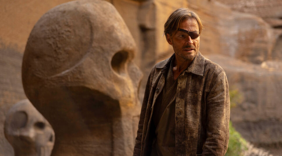 In „Die drei ??? – Toteninsel“ steht Professor Phoenix, gespielt von Andreas Pietschmann, vor monumentalen Steinskulpturen in Form von Totenschädeln. Mit Augenklappe, wettergegerbter Kleidung und ernstem Blick wirkt er entschlossen und geheimnisvoll.