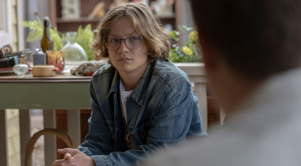 Bob Andrews (Levi Brandl) ist in einer Szene aus "Die drei ??? und der Karpatenhund" zu sehen. Er trägt eine Brille und eine Jeansjacke und sitzt in einem Raum mit Pflanzen und dekorativen Gegenständen im Hintergrund. Sein Gesichtsausdruck wirkt nachdenklich und fokussiert.