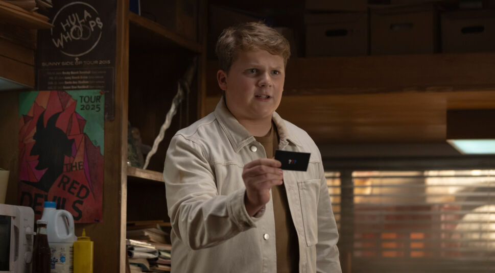 Justus Jonas (gespielt von Julius Weckauf) in einer Szene aus "Die drei ??? und der Karpatenhund". Er trägt eine helle Jacke und hält eine Visitenkarte mit dem "???"-Logo in der Hand.