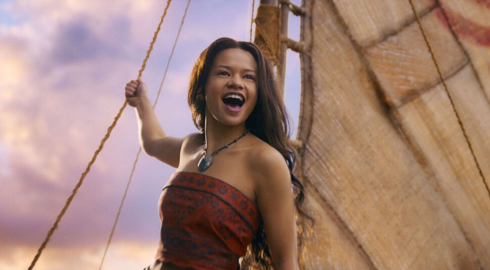 In dieser Szene aus der Vaiana-Realverfilmung ist Catherine Laga‘aia als Vaiana zu sehen. Sie steht auf einem traditionellen Segelboot, hält sich an einem Seil fest und blickt freudestrahlend in die Ferne.