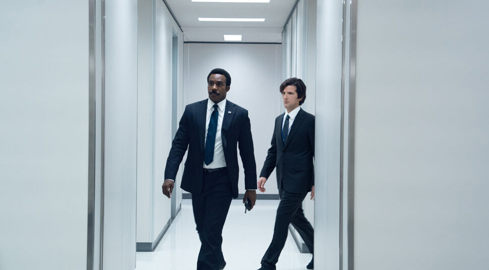 Auf dem Bild zu Severance Staffel 3 sind rechts Mark Scout, gespielt von Adam Scott, und links Seth Milchick, gespielt von Tramell Tillman, in einem sterilen Büroflur zu sehen. Beide tragen dunkle Anzüge und ernste Gesichtsausdrücke. Milchick geht entschlossen mit einem Objekt in der Hand voran, während Scout ihm aufmerksam folgt. Die weiße Umgebung unterstreicht die kühle, kontrollierte Atmosphäre der Szene. Das Bild stammt aus Staffel 2 von Severance.