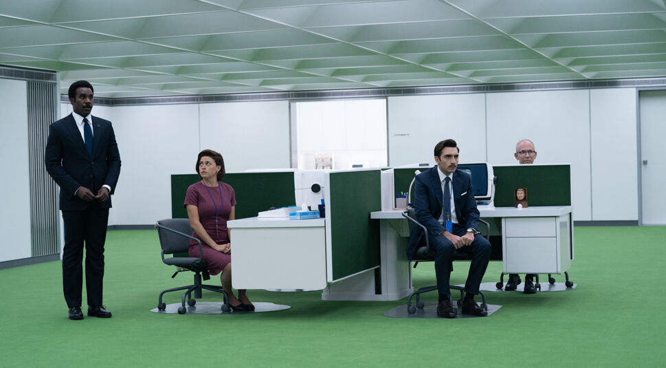 Auf dem Bild aus Staffel 2 von Severance sind von links nach rechts Seth Milchick, gespielt von Tramell Tillman, Gwendolyn Y., gespielt von Alia Shawkat, Dario Rossi, gespielt von Stefano Carrannante, und Mark Wilkins, gespielt von Bob Balaban, in einem sterilen Büro mit grünem Teppichboden zu sehen. Milchick steht förmlich, während die anderen an weißen Schreibtischen sitzen. Die minimalistische Büroeinrichtung und die kühle Atmosphäre betonen die kontrollierte Arbeitsumgebung. Severance Staffel 3 ist bereits bestätigt.