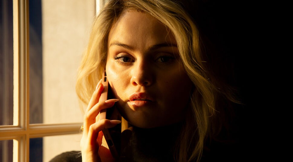 Auf dem Bild aus Emilia Pérez ist Selena Gomez in der Rolle von Jessi zu sehen. Sie hält ein Smartphone ans Ohr und blickt nachdenklich und angespannt in die Ferne. Das warme Licht von einem Fenster beleuchtet ihr Gesicht, während der Rest der Szene in Schatten gehüllt ist. Die Stimmung wirkt geheimnisvoll und dramatisch. Emilia Pérez streamen ermöglicht es, diesen intensiven Moment mitzuerleben.