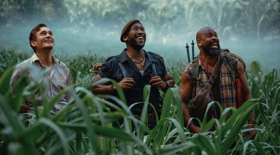 Auf dem Bild siehst Du eine Szene aus Jurassic World: Die Wiedergeburt. Von links nach rechts sind Rupert Friend als Martin Krebs, Mahershala Ali als Duncan Kincaid und Bechir Sylvain als Leclerc zu sehen. Die drei Männer stehen inmitten eines dichten, grünen Feldes und blicken lachend in den Himmel.