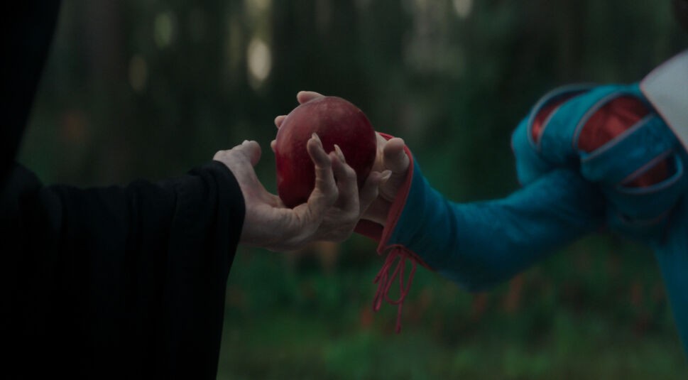 Auf dem Bild aus Schneewittchen (2025) ist der ikonische Moment dargestellt, in dem Schneewittchen den verzauberten Apfel von der verkleideten bösen Königin entgegennimmt. Die Szene ist dunkel und atmosphärisch gehalten, mit Fokus auf die Hände – eine in einem schwarzen Umhang, die andere in einem detaillierten blauen Kleid mit roten Akzenten. Im Vergleich zur stilisierten Animation von Schneewittchen und die sieben Zwerge (1937) wirkt diese Neuinterpretation realistischer und düsterer, was die moderne Inszenierung unterstreicht.