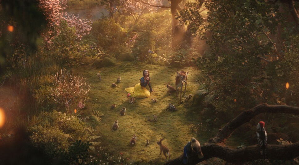 Auf dem Bild aus Schneewittchen (2025) sitzt Rachel Zegler als Schneewittchen inmitten eines idyllischen Waldes, umgeben von Rehen, Kaninchen und Vögeln. Sonnenlicht fällt durch die Bäume und verleiht der Szene eine märchenhafte, fast magische Atmosphäre. Ihr Kleid in Blau, Rot und Gelb erinnert an das klassische Design des Originalfilms Schneewittchen und die sieben Zwerge (1937), während die realistische CGI-Umgebung die moderne Neuinterpretation betont. Dieses Bild zeigt den visuellen Wandel vom handgezeichneten Animationsstil zur cineastischen Realverfilmung.