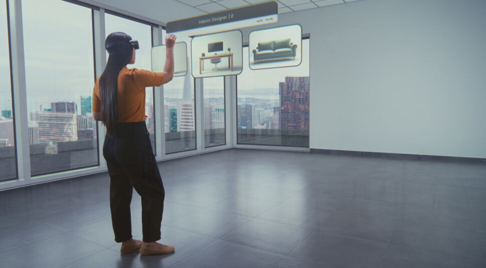 Auf dem Bild zum Thema Was ist Extended Reality steht eine Frau mit XR-Headset in einem leeren Raum mit Panoramafenstern. Vor ihr schwebt ein digitales Interface mit Möbelstücken, das sie per Handbewegung bedient. Die Szene zeigt den Einsatz von Extended Reality für virtuelle Innenraumplanung und veranschaulicht die Verschmelzung von Realität und digitaler Gestaltung.