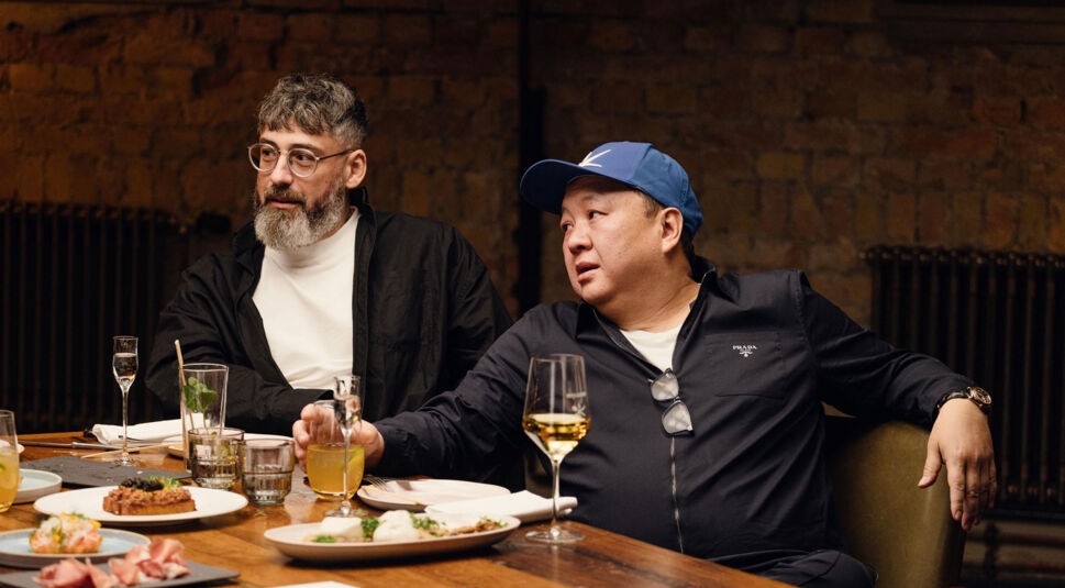 In dieser Szene aus Kitchen Impossible Staffel 10 sitzen Sido und The Duc Ngo nebeneinander an einem rustikalen Holztisch.