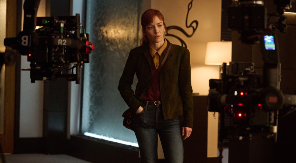 Im Bild zur Serie Nur für Dein Leben ist Britt Lower als Rachel Mills in einem modern eingerichteten Innenraum zu sehen. Sie steht zwischen zwei Filmkameras, trägt eine dunkelgrüne Jacke und schaut konzentriert zur Seite. Die Szene wirkt ruhig und nachdenklich, mit warmem Licht und stilisiertem Hintergrunddesign.