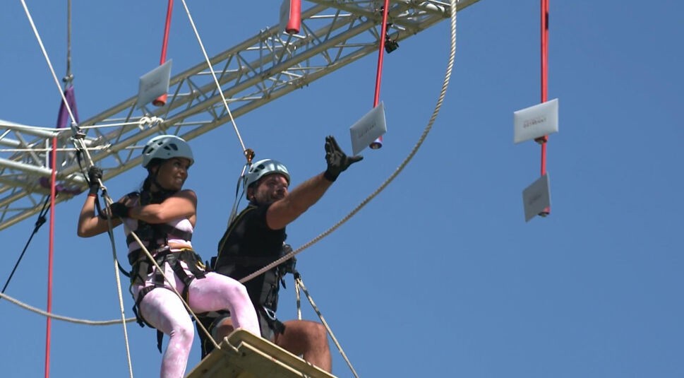 In einer actionreichen Challenge der ersten Staffel von „Prominent getrennt“ (2022) kämpfen Meike Emonts und Marcus Muth gemeinsam in luftiger Höhe. Beide tragen Helme und Sicherheitsgurte, während Marcus nach einem Umschlag greift. Die Szene zeigt den Einsatz der späteren „Prominent getrennt-Gewinner:innen“ unter strahlend blauem Himmel.