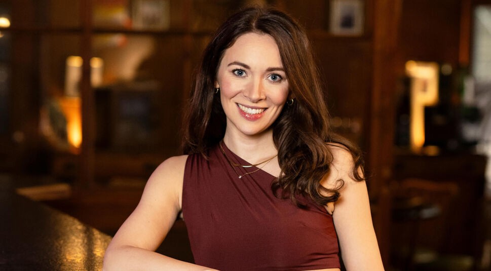 Eine Kandidatin von Die Bachelors 2025 lehnt an einem Tresen, trägt ein weinrotes, schulterfreies Top und hat lange, braune Haare.