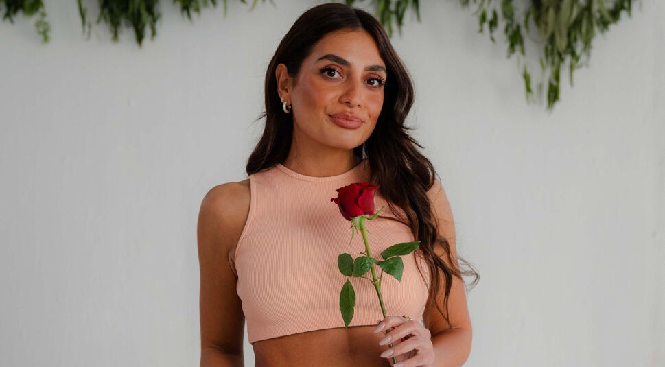 Eine Kandidatin von Die Bachelors 2025 hält eine rote Rose in der Hand und trägt ein bauchfreies, apricotfarbenes Top vor weißem Hintergrund.